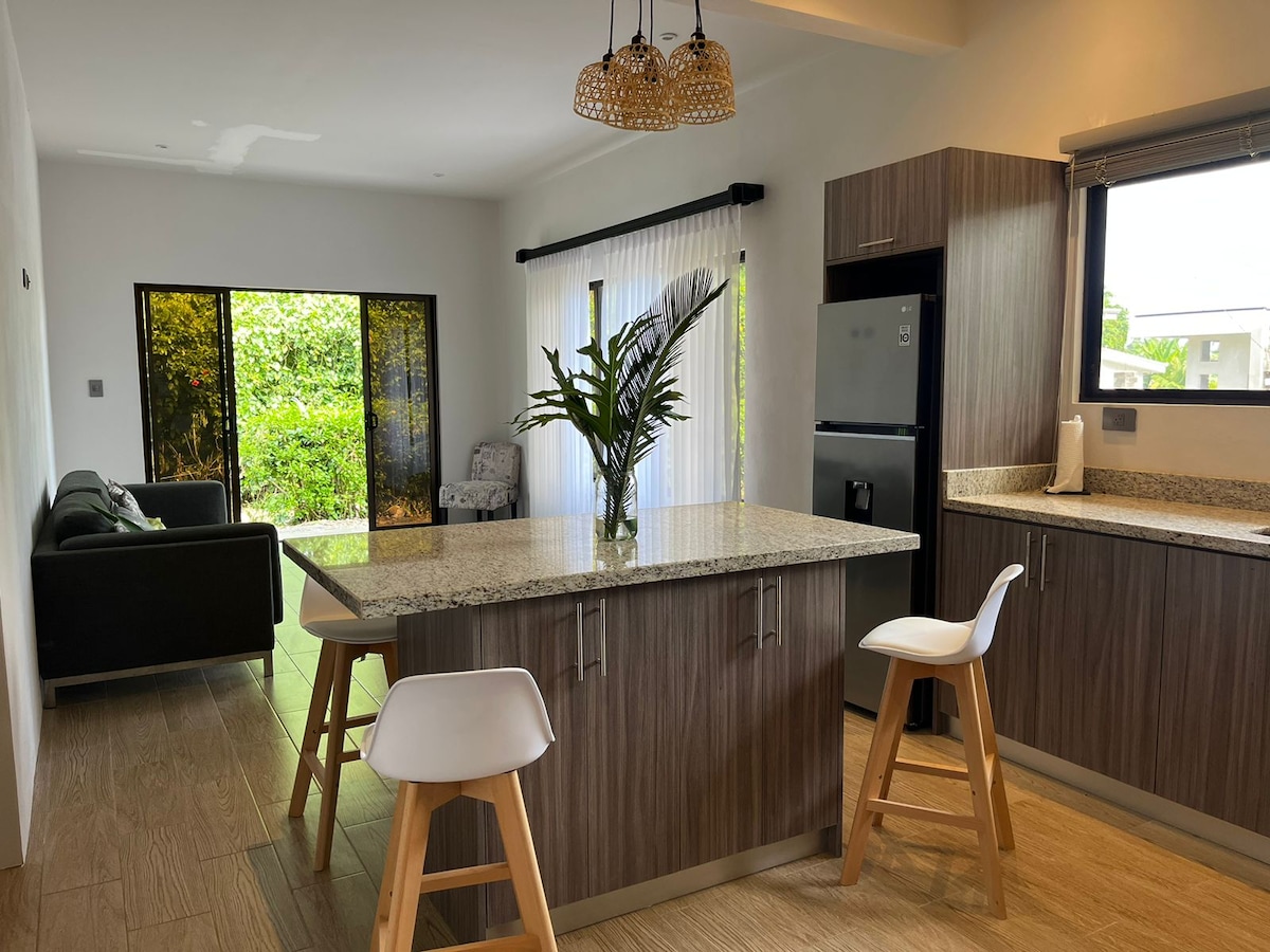 The modern kitchen features a central island with two wooden stools, gray cabinets, and a refrigerator. A window provides views of the outdoors, where lush greenery is visible. A small seating area with a couch is seen in the adjacent space.