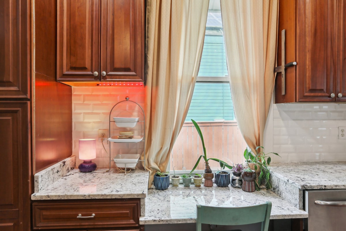 The kitchen features rich wooden cabinets and a granite countertop. Natural light filters through a large window adorned with soft curtains. A small side table displays bowls and plants, contributing to a pleasant atmosphere, while a subtle illumination highlights the countertop's textures.