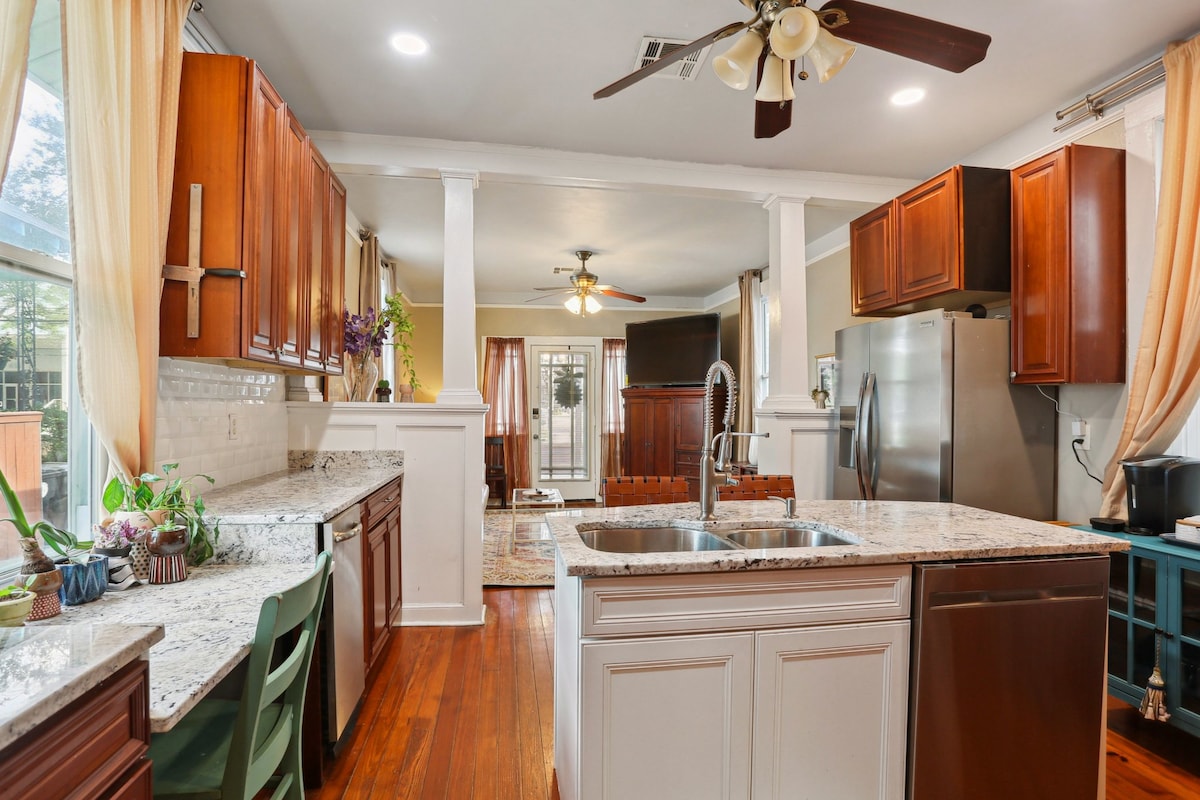 A spacious kitchen features dark wood cabinetry and granite countertops. Natural light floods the area through large windows, enhancing the inviting atmosphere. A central island with a double sink is surrounded by a dining table and modern appliances, contributing to a functional cooking space.