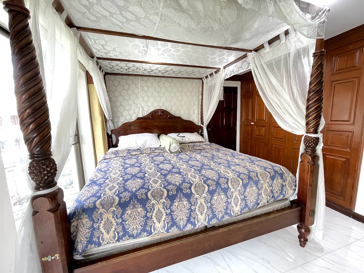 A four-poster bed, framed by sheer curtains, occupies the center of the room. The bedding features a blue and gold patterned cover, while a decorative pillow rests against the headboard. Natural light fills the space, highlighting the sleek tile flooring and wooden accents in the background.
