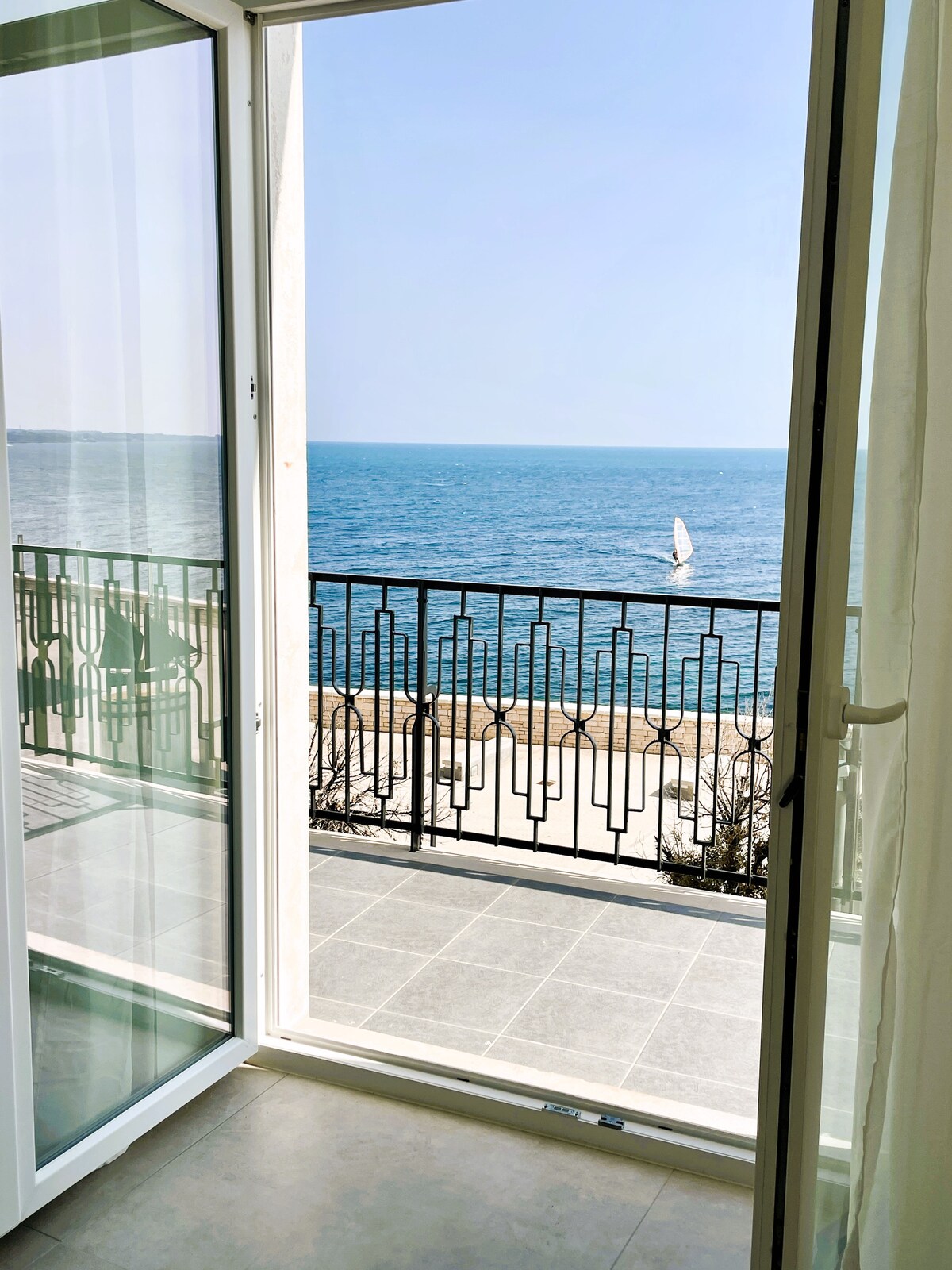 A large glass door opens to a sea view, revealing a spacious balcony with a decorative railing. The calm water reflects the clear sky, and a sailboat is visible in the distance, enhancing the tranquil seaside setting.