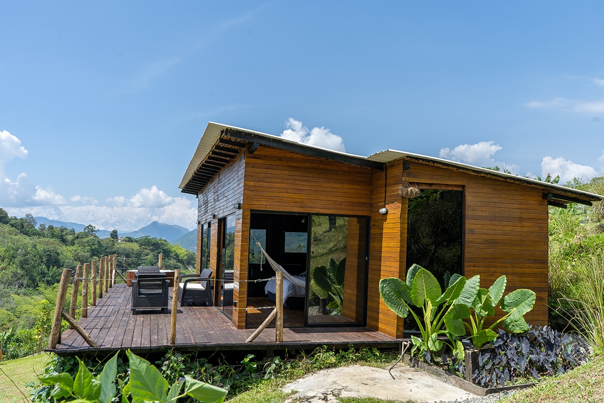 A wooden cabin is set against a backdrop of rolling hills and blue skies. Expansive glass doors open onto a spacious deck surrounded by lush greenery. The outdoor seating area invites relaxation, complementing the serene natural landscape.