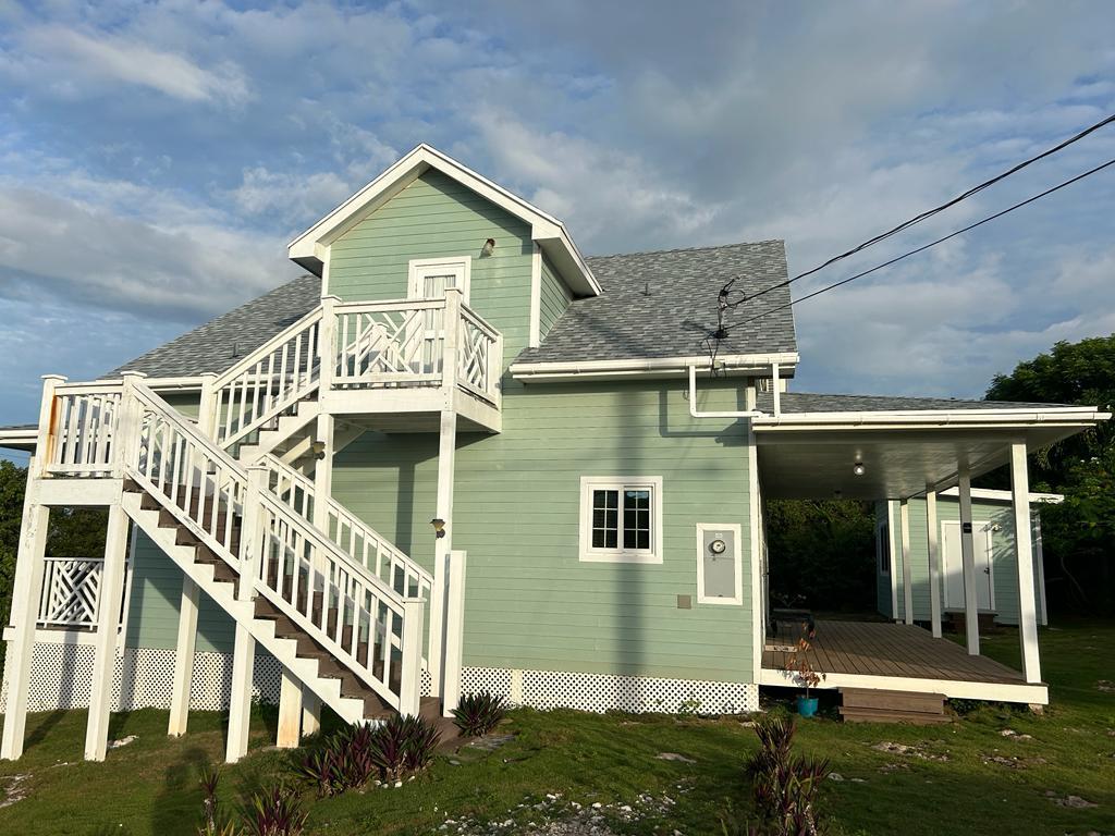 A two-story house features a light green exterior and a tiered staircase leading to the entrance. A spacious covered porch is visible on the side, and is surrounded by well-maintained landscaping, highlighting the peaceful setting.