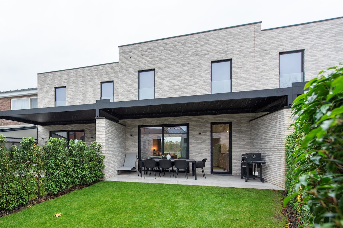 A spacious outdoor area features a covered patio with dining table and chairs, accompanied by a barbecue grill. Lush greenery frames the lawn, while large windows and glass doors create a seamless transition between indoor and outdoor spaces.