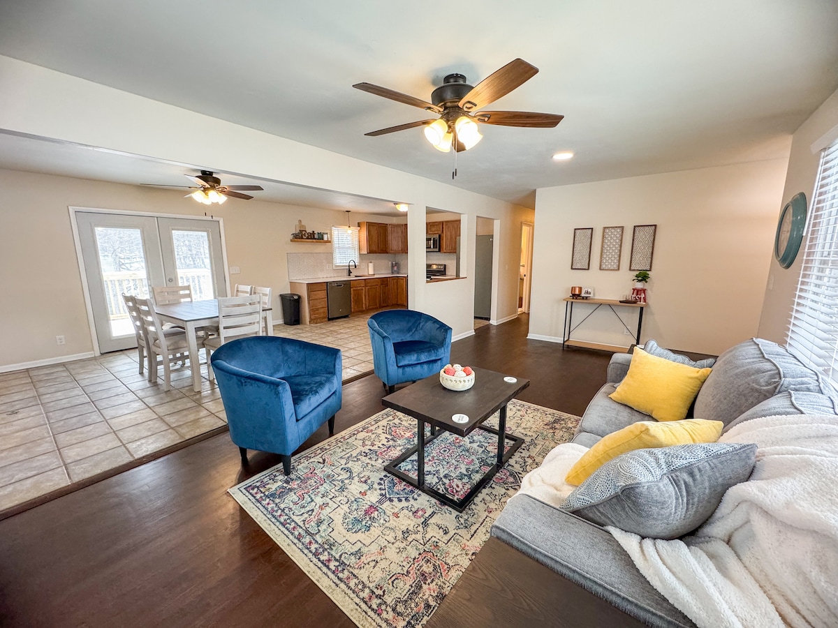 A spacious living room features a comfortable gray sofa with a yellow accent pillow, two blue armchairs, and a decorative area rug. A coffee table is placed in the center. Light streaming through the large windows enhances the warm ambiance, while the open layout connects to a dining area and kitchen.