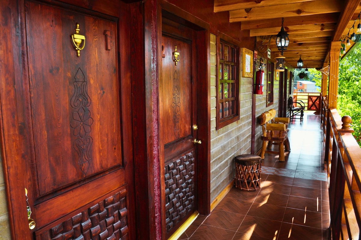 A warm wooden walkway is lined with intricately carved doors leading to the rooms. Soft lighting is provided by decorative lanterns, enhancing the natural tones of the wood. The surrounding green landscape is visible through large windows, creating a tranquil ambiance.