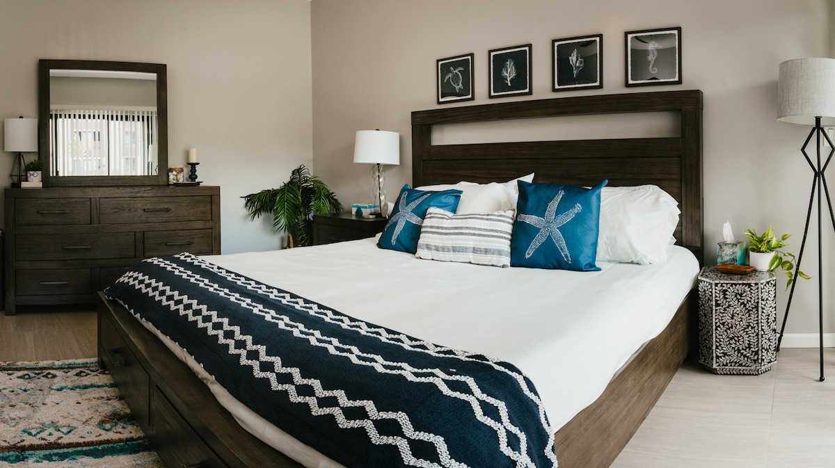 A spacious bedroom features a large bed with neutral bedding accented by decorative pillows in shades of blue. Framed artwork adorns the wall above, and a stylish dresser and mirror are situated nearby. A floor lamp and potted plant add a touch of warmth to the space.