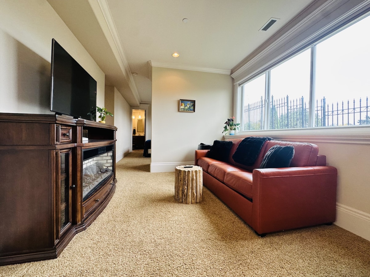 A cozy living area features a red leather sofa positioned in front of a television and a wooden entertainment unit. Natural light filters in through large windows, enhancing the warm tone of the space. A small rustic table made from a tree stump is placed beside the sofa.