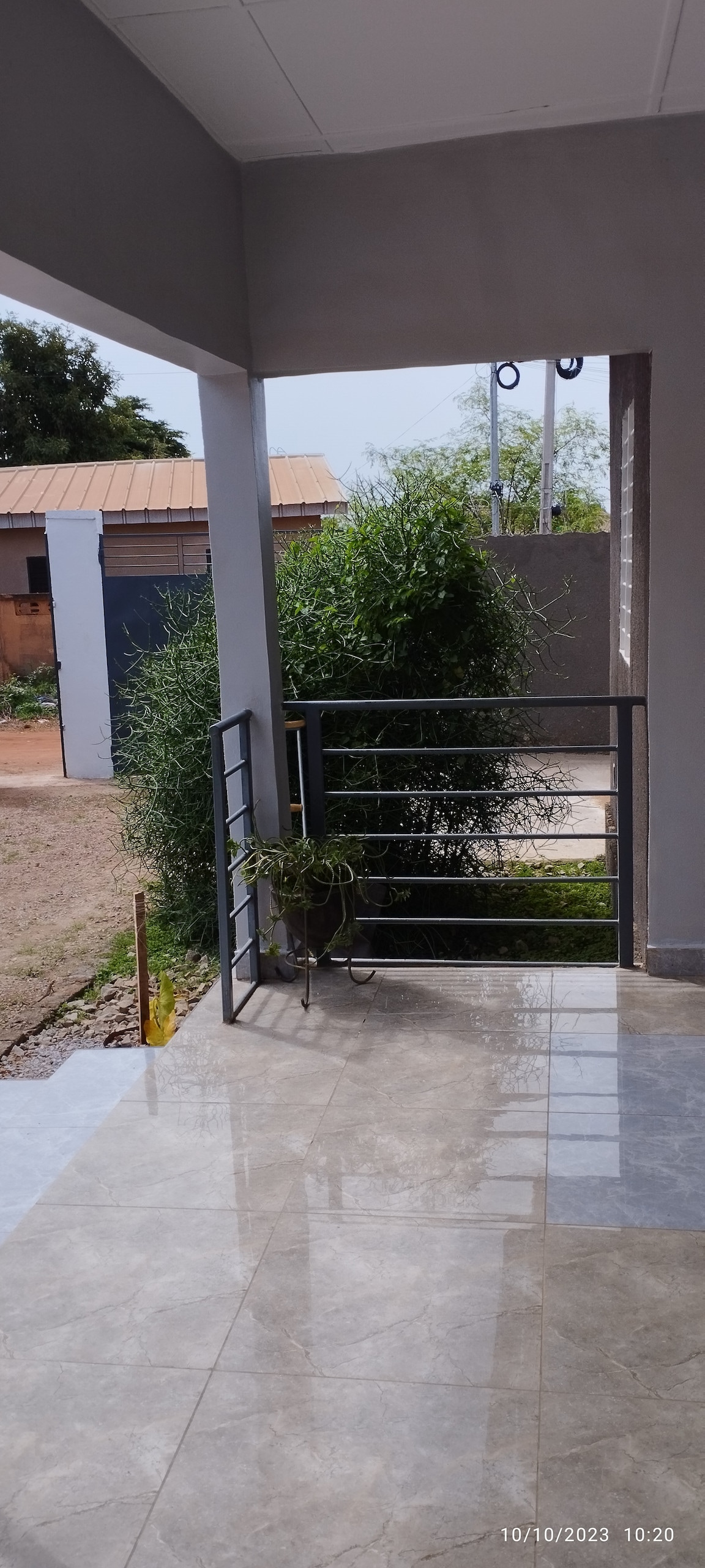 A covered entryway features polished tile flooring, with a railing that leads to an outdoor area. Lush greenery is visible beyond a low fence, while the surrounding walls are painted in a soft gray hue, creating an inviting atmosphere.