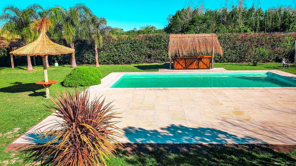 A tranquil outdoor space features a clear swimming pool surrounded by lush greenery. A thatched-roof structure provides shade nearby, while a well-maintained lawn and varied plant life enhance the serene setting.
