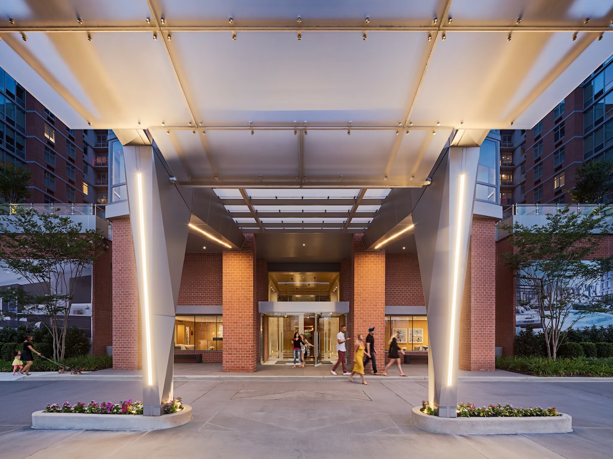 An impressive entrance is showcased, featuring modern architectural elements with metal and glass structures. Soft lighting highlights the entrance, while people walk along a paved pathway. Lush greenery and carefully arranged flowers are visible along the edges, enhancing the welcoming appeal.