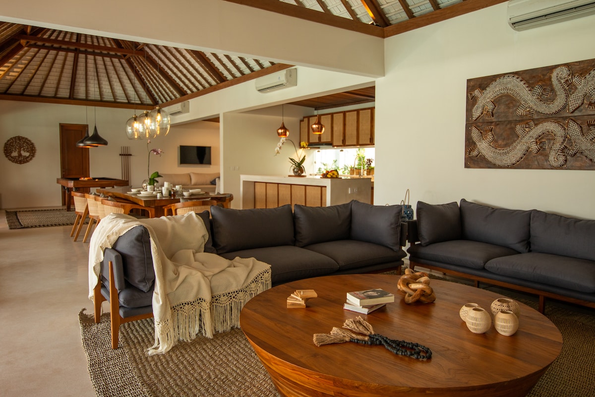 An open living area features a large, round wooden table surrounded by comfortable seating. Natural light floods the space, highlighting the textured decor and neutral colors. A cozy throw and decorative objects add warmth, while a modern kitchen is visible in the background.