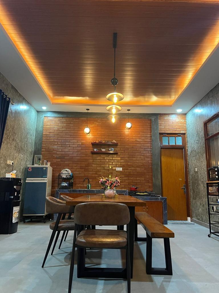 A modern kitchen area features a large wooden table surrounded by four brown chairs. Pendant lights hang from a warm wood ceiling, illuminating the space. A brick wall adds texture, while a refrigerator and other kitchen appliances are neatly arranged along one side.