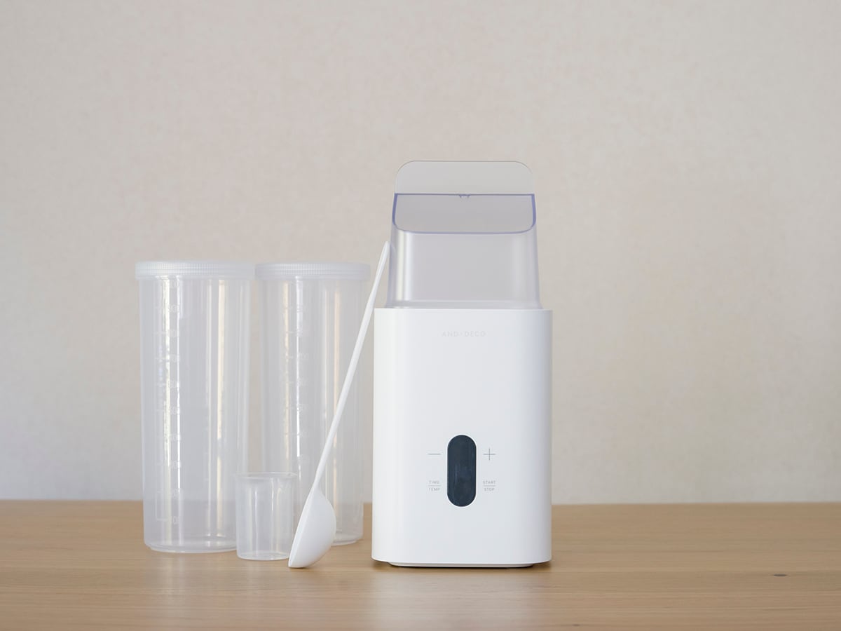 A sleek white appliance is presented on a wooden surface. Two clear containers are positioned beside it, along with a minimalist stirring tool. The design emphasizes clean lines and functionality, suitable for kitchen use.