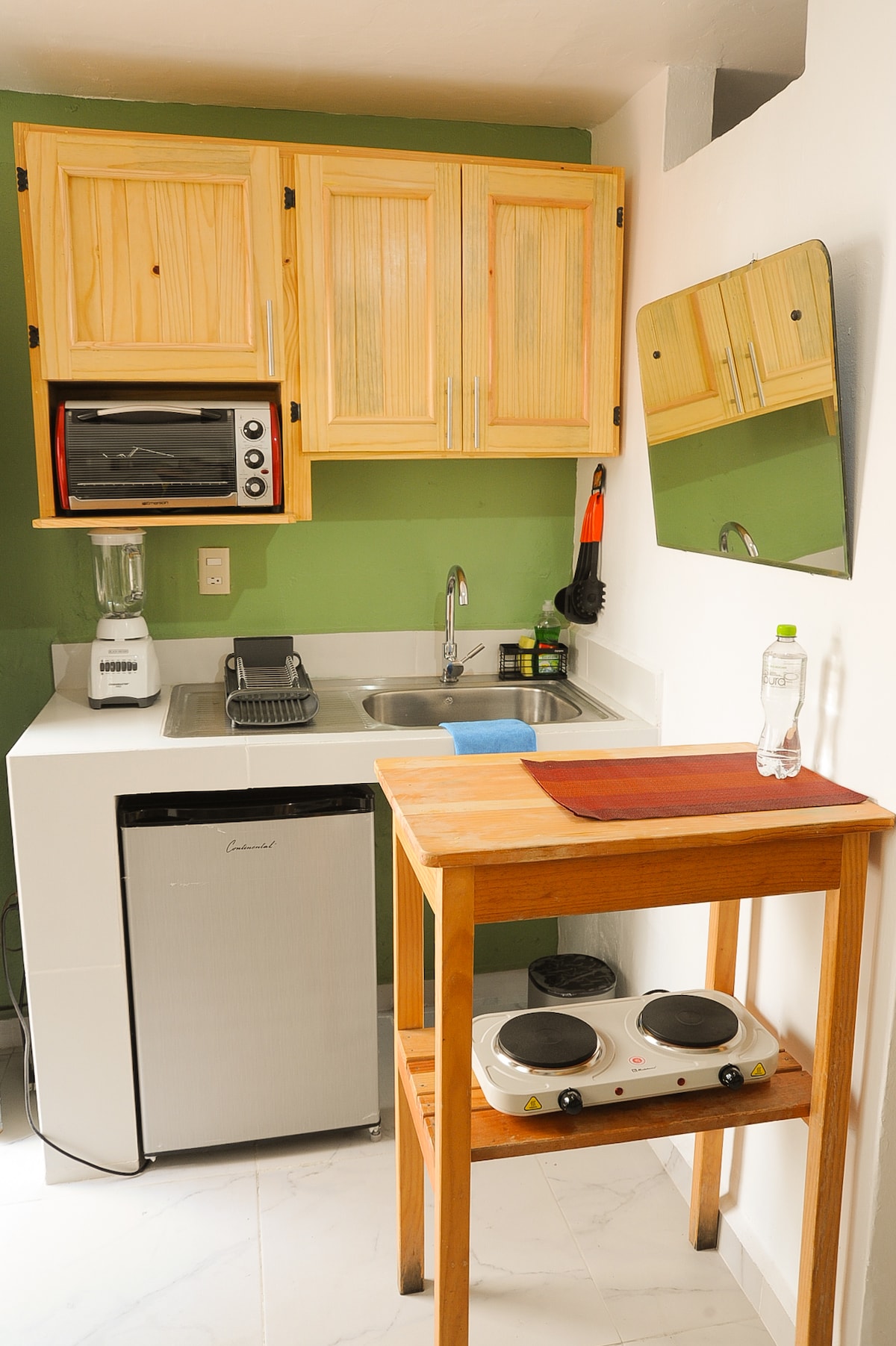 A compact kitchenette is equipped with wooden cabinets, a microwave, and a blender. A small refrigerator is placed alongside a sink. A wooden table holds two hot plates, providing additional cooking options. The green wall adds a fresh touch to the functional space.