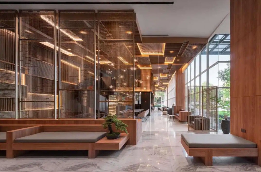 A modern lobby features a mix of wood and glass elements, providing a spacious and inviting atmosphere. Seating areas with wooden benches and cushions are arranged alongside decorative plants. Natural light streams in through large windows, complemented by soft ambient lighting overhead.