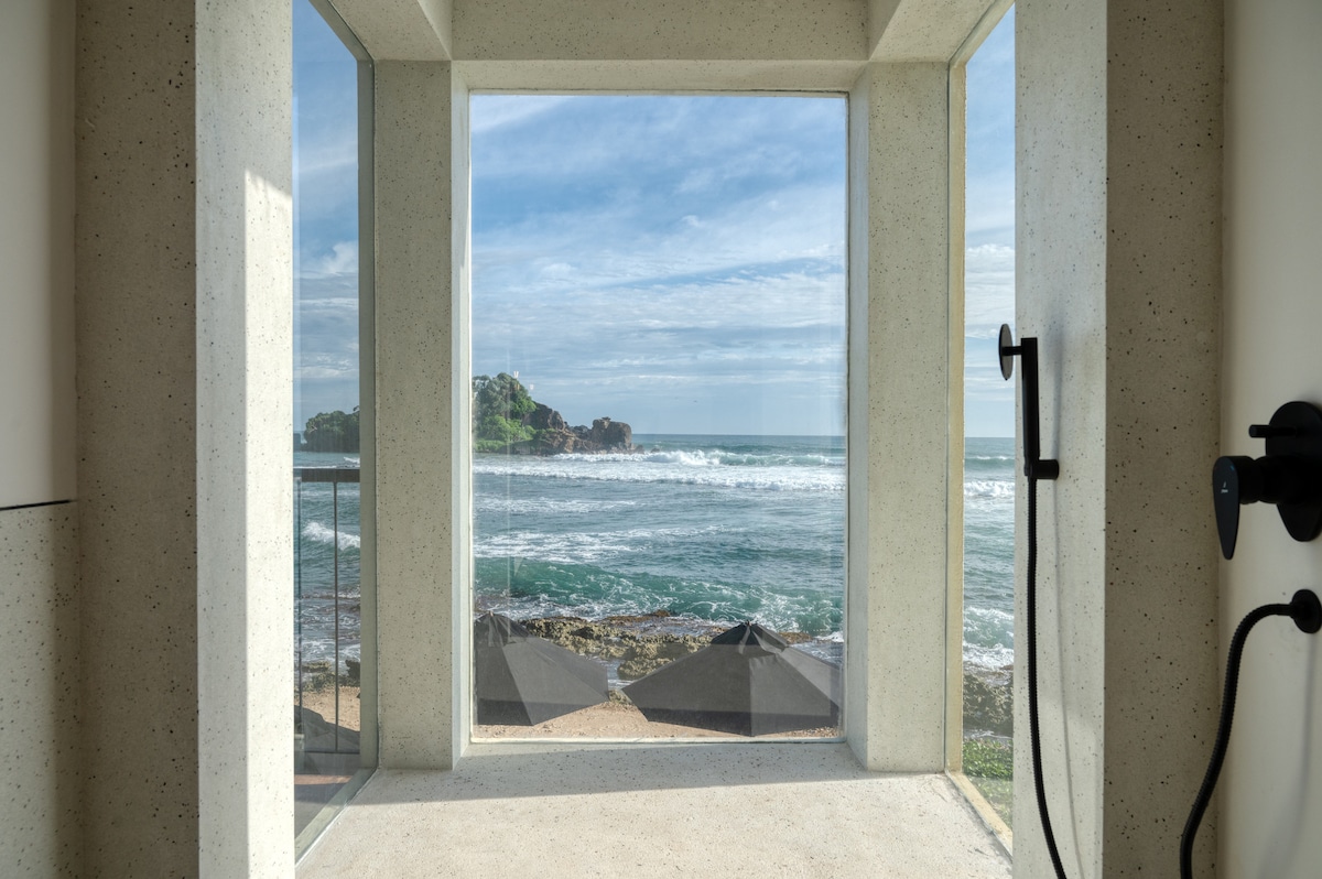 A window frames a scenic view of the ocean waves and rocky shoreline, with lounge umbrellas visible on the beach below. The minimalist design features concrete walls and a sleek shower fixture, contributing to the overall modern aesthetic.
