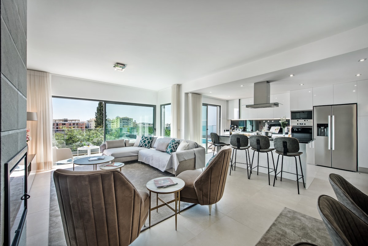 A spacious living area is presented with large windows offering views of the surrounding area. Modern furnishings include a comfortable sectional sofa and sleek coffee tables. A fully equipped kitchen with bar seating is visible in the background, creating an inviting space for gatherings.