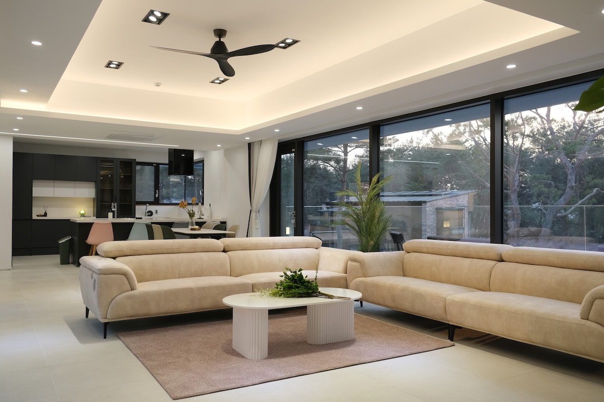 A spacious living area features a soft-toned, sectional sofa arranged around a modern coffee table. Large glass windows allow natural light to fill the room, providing views of the surrounding greenery. A ceiling fan circulates air above, enhancing the comfortable atmosphere.