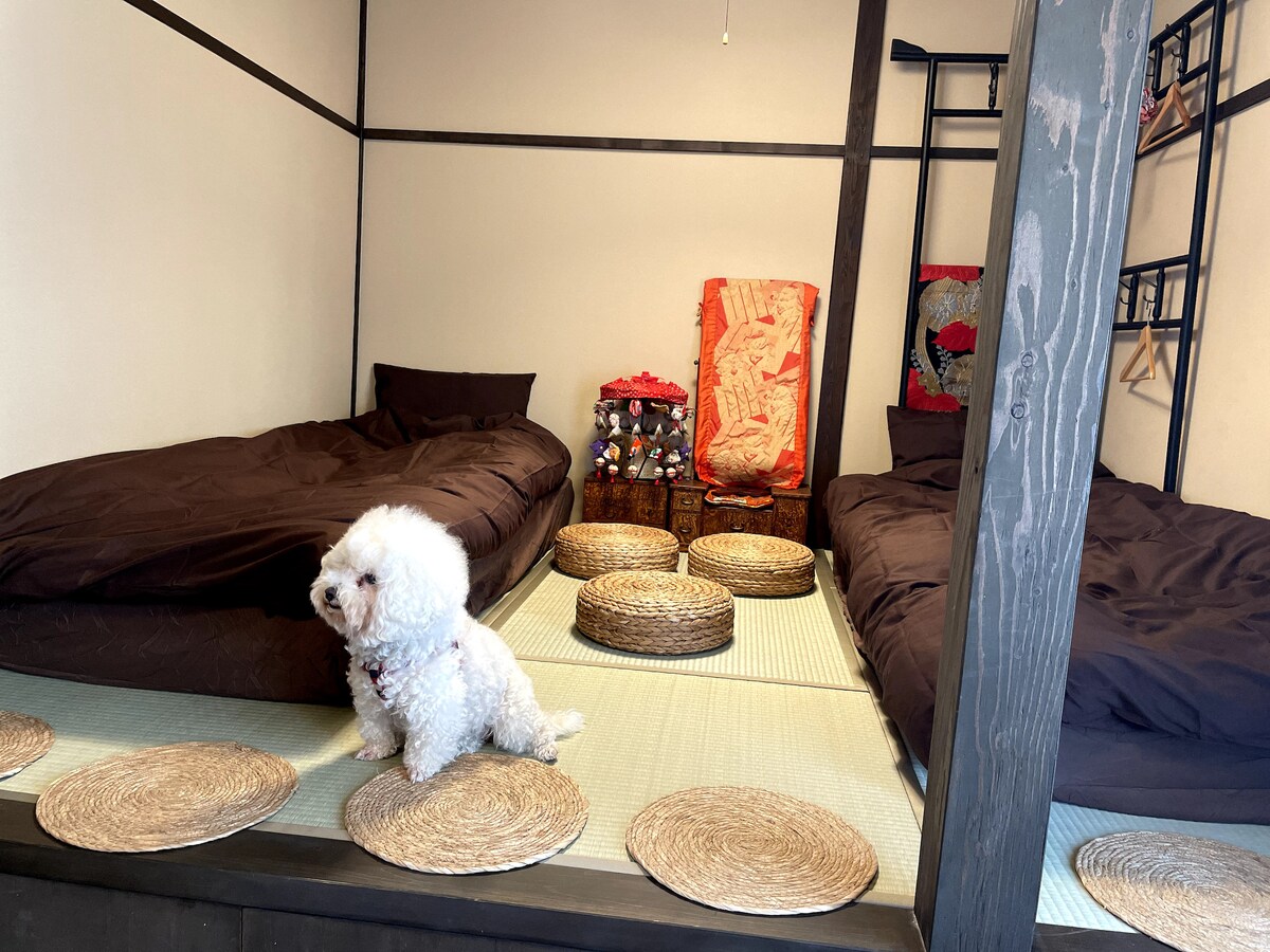 A serene sleeping area is showcased with two single beds dressed in dark bedding. Round woven mats are placed on tatami flooring, providing an inviting space. A small dog sits nearby, adding a lively touch to the tranquil setting, complemented by a decorative chair in the background.