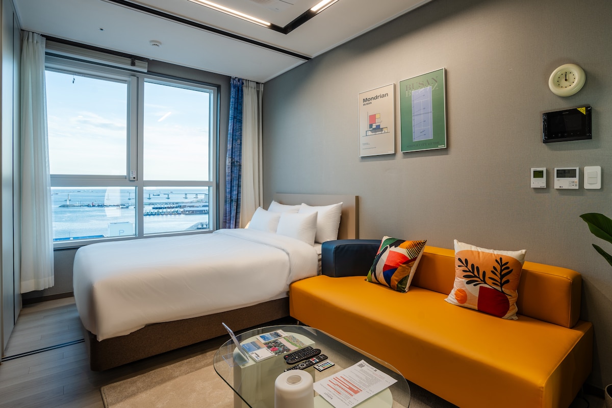 A bright hotel room features a spacious bed dressed in white linens, complemented by a vibrant orange sofa with decorative pillows. Large windows offer views of the ocean, and artwork adorns the walls. Modern amenities include a digital panel and comfortable furnishings.
