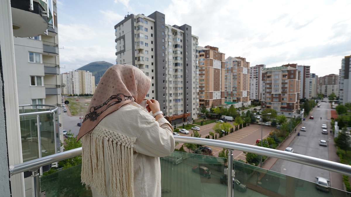 A view from a balcony captures surrounding buildings and a tree-lined street. A figure in a headscarf is seen, facing the outdoors, with a soft breeze visible in the environment. The background includes multi-story residences and distant hills under a mostly cloudy sky.