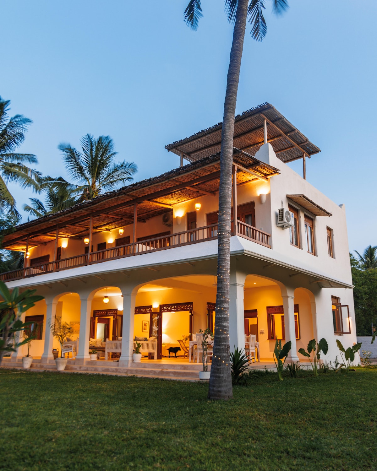 The villa's exterior showcases a modern architectural design with multiple levels. A spacious veranda with wooden railings overlooks the lush garden, while palm trees frame the property. Warm lighting creates an inviting ambiance as evening approaches, enhancing the tranquil setting.
