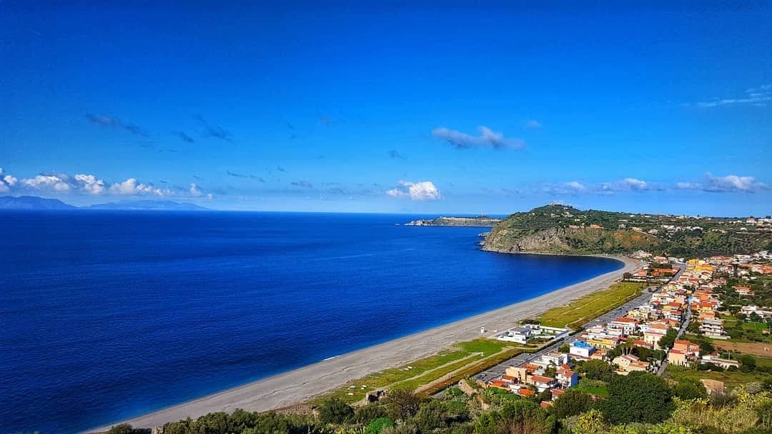 A panoramic view of a coastal landscape highlights a stretch of sandy beach lined with a small town. Clear blue skies merge with the calm sea, while hills frame the scene. The beach area extends gently, with patches of greenery visible along the shoreline.