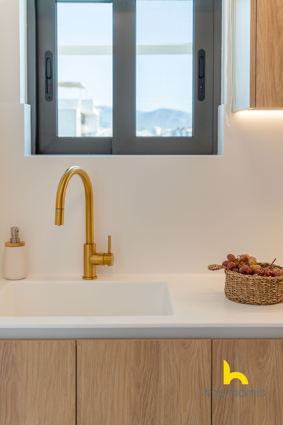 A modern kitchen space showcases a stylish gold faucet above a sleek, white sink. Natural light enters through a window framed by minimalist black accents. A basket filled with grapes is placed on the countertop, complementing the warm wooden cabinetry below.