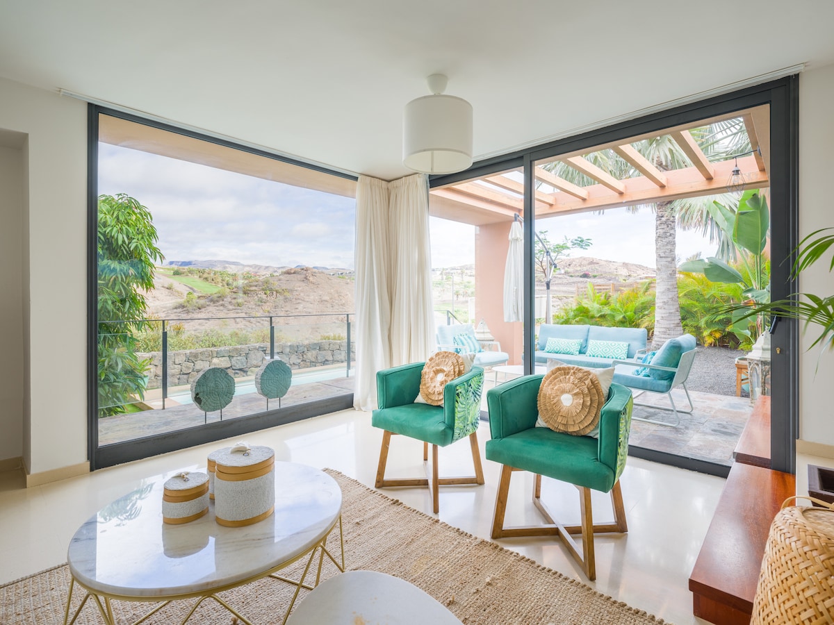 A bright living area features large glass doors that open to a patio with lush views. Two teal armchairs are positioned near a round coffee table, complemented by a natural fiber rug. Potted plants add greenery to the space, creating an inviting ambiance.