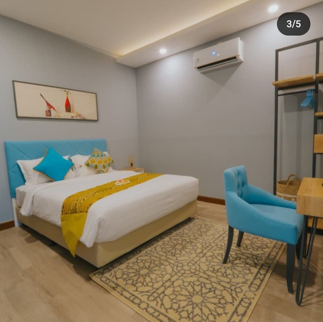 A well-appointed bedroom features a spacious bed with white linens and decorative pillows. A vibrant blue chair complements a wooden desk. A patterned rug is positioned on the light-colored floor, while a piece of artwork hangs above the bed, adding a touch of color to the neutral walls.