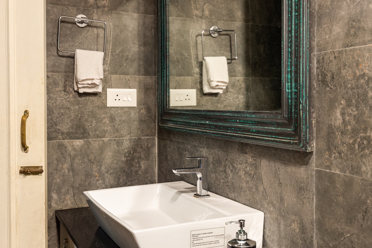A modern bathroom is shown, featuring a sleek white sink positioned on a dark countertop. A large mirror with a decorative frame enhances the space. Two neatly hung towels are placed on a bar above the sink, set against a textured gray tiled wall.