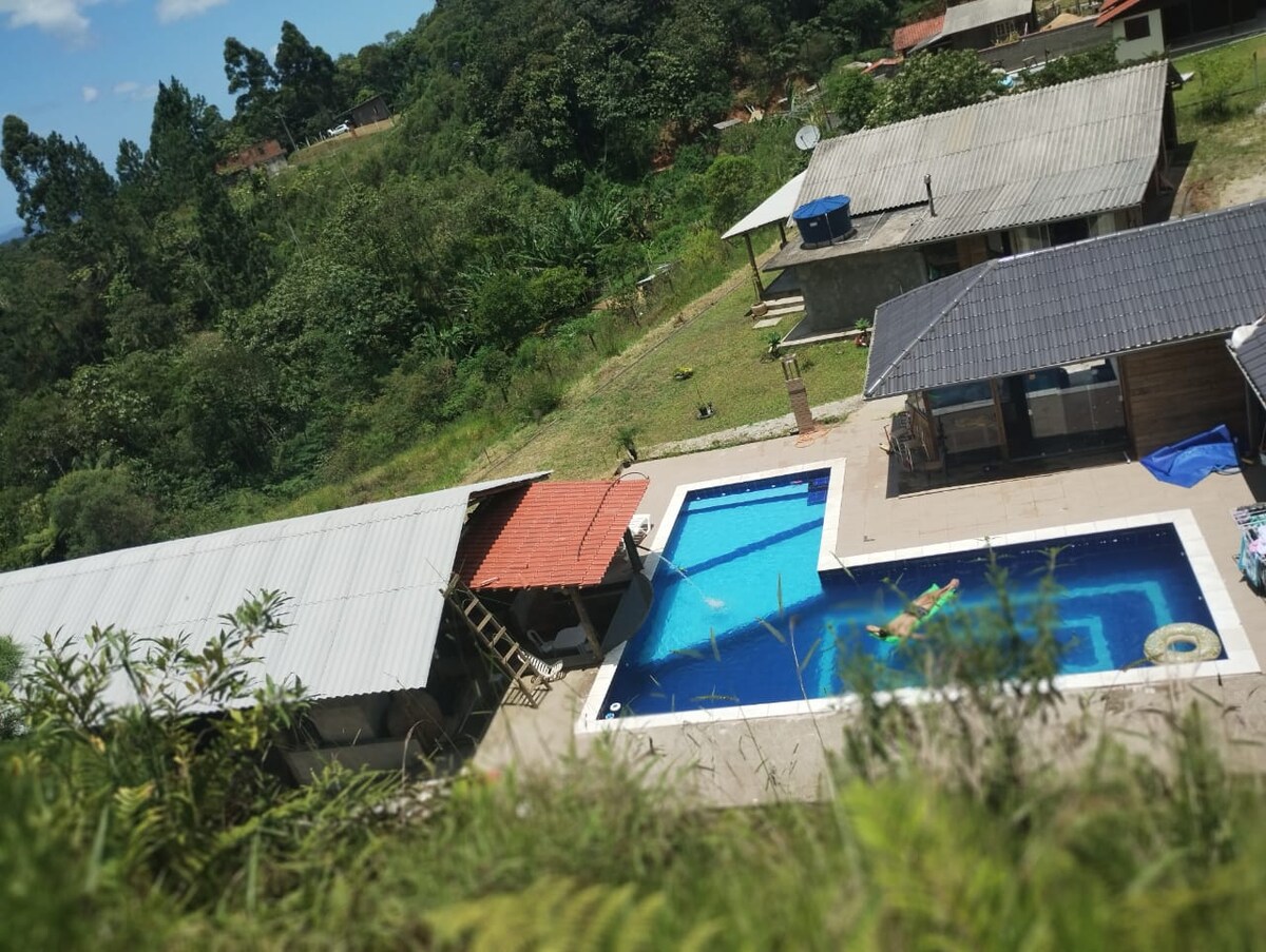 An expansive outdoor area is visible, featuring a large swimming pool surrounded by tiled decking. A shaded patio structure and other buildings are situated nearby, with lush greenery and a hilly landscape in the background, emphasizing the property's tranquil natural setting.