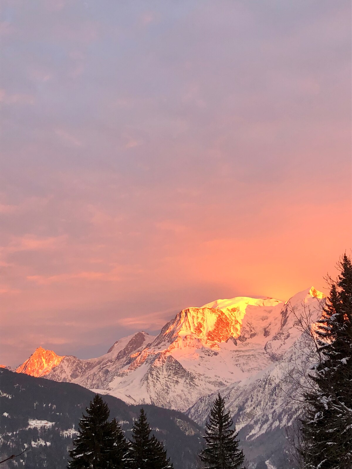 Snow-capped mountains are illuminated by the warm glow of sunrise, contrasting with a soft pink and orange sky. Evergreen trees frame the scene, enhancing the natural beauty of the alpine landscape.