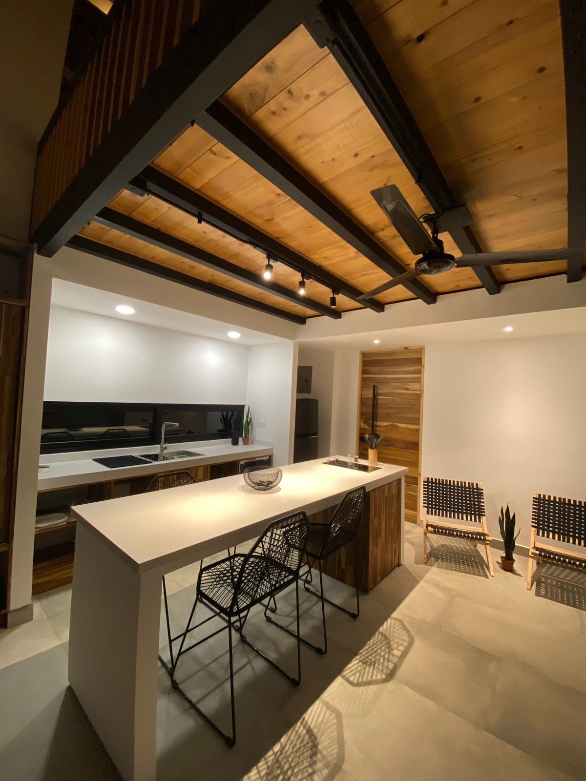 A modern kitchen space features a sleek white island with black wire stools. Warm wood beams line the ceiling above, and the walls are painted in bright white. Subtle lighting enhances the clean design, while potted plants add a touch of nature to the area.