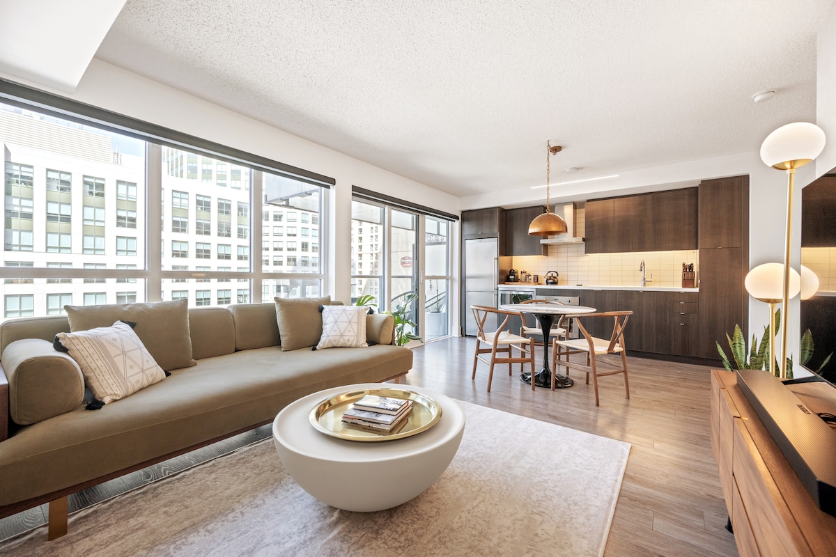A modern living space showcases a spacious, L-shaped sofa arranged around a round coffee table. Large windows provide natural light and views of the city. A dining area with four chairs is visible, along with a well-equipped kitchen featuring sleek wooden cabinetry and pendant lighting.