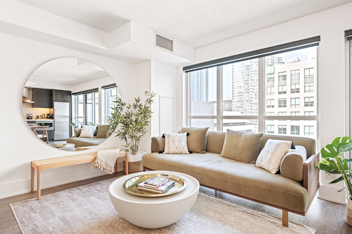 A spacious living area features a comfortable sofa near large windows, allowing natural light to fill the room. A round mirror reflects the stylish decor, while a coffee table with magazines adds to the inviting atmosphere. A potted plant and decorative elements enhance the space.