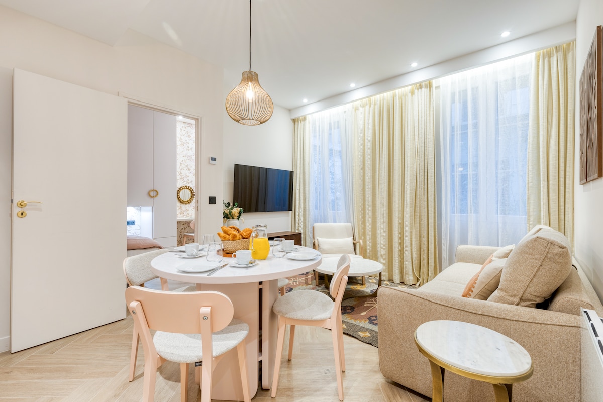 A dining area features a round table surrounded by four light-colored chairs. Natural light enters through sheer curtains, illuminating a cozy sofa nearby. A television is mounted on the wall, contributing to the inviting space. A door leads to another room, enhancing the apartment's layout.
