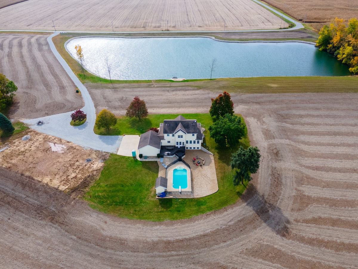 An aerial view captures a country retreat surrounded by farmland. A spacious yard includes an in-ground pool and a stocked pond, with winding pathways leading to the home. Colorful trees are positioned nearby, showcasing seasonal foliage against the expansive landscape.