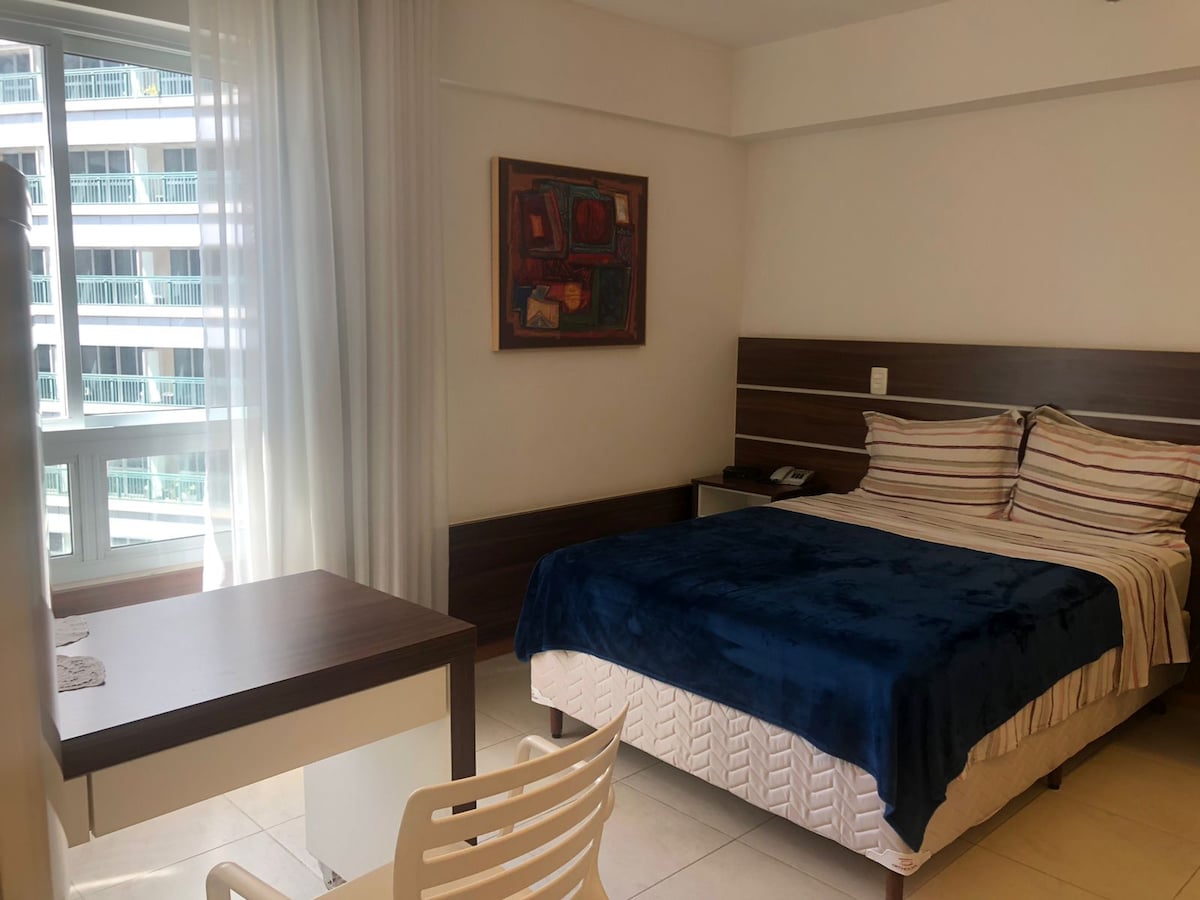 A bright and airy room is presented, featuring a spacious bed covered with a blue blanket. A desk and chair are positioned nearby, and large windows allow natural light to filter through sheer curtains. Abstract artwork adorns the wall above the bed.