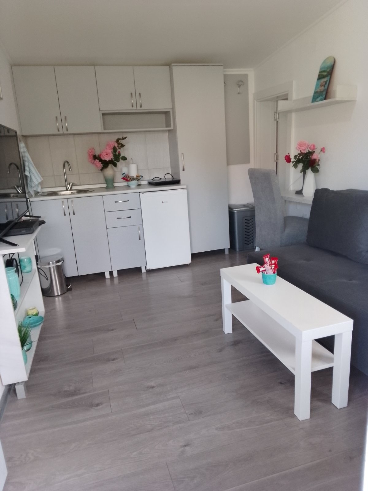 A bright and functional kitchenette displays white cabinets and a countertop. A cozy seating area features a gray sofa next to a white coffee table. Decorative flowers are arranged on the counter, contributing to the space’s fresh ambiance.
