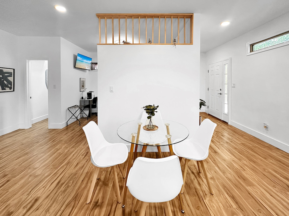 A dining area is presented with a round glass table surrounded by four white chairs. Natural light enters through nearby windows, illuminating the light wood flooring. A small plant decorates the table, while a workspace is subtly visible in the background.