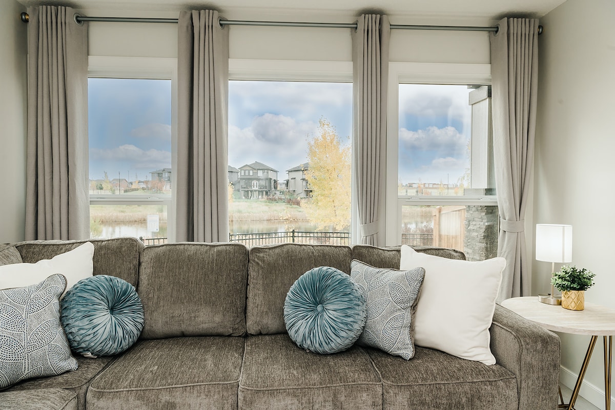 A comfortable sectional sofa is adorned with patterned and solid throw pillows. Natural light enters through large windows dressed with flowing curtains, framing views of a serene pond and surrounding residences.