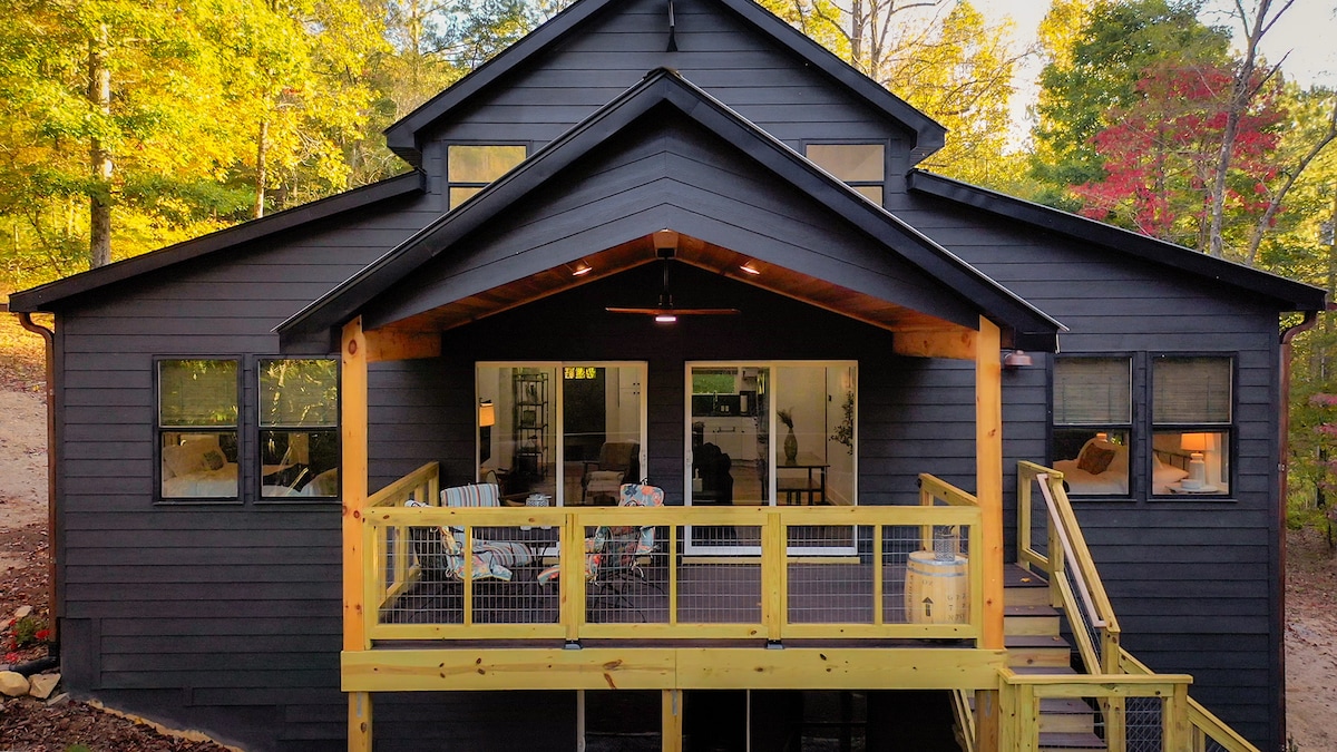 A modern barndominium-style cabin is presented with a dark exterior, surrounded by trees. The spacious deck features a railing and steps leading to a cozy interior, where large windows allow natural light to fill the living area.