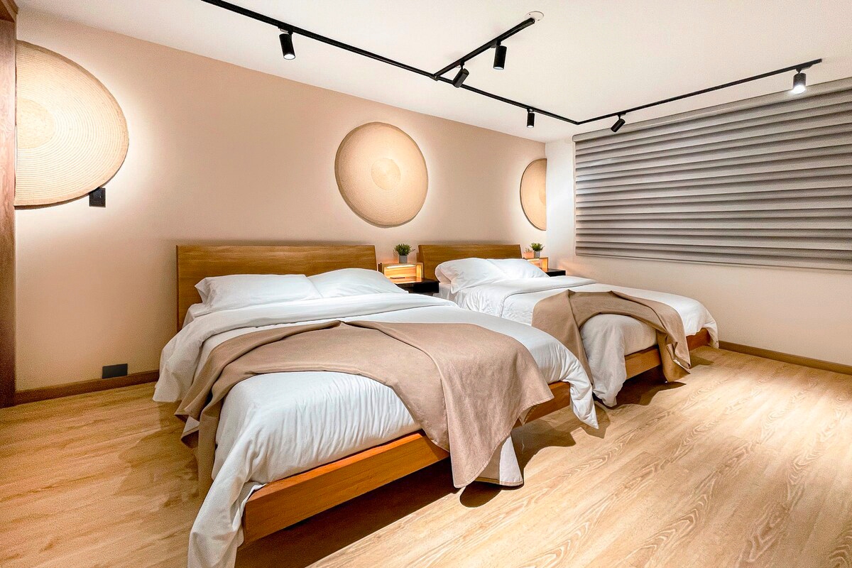 The sleeping area features two spacious beds with neutral-toned bedding and soft brown throws draped over them. Natural light filters through the window coverings, highlighting the wooden floors and wall decor. The room is illuminated by overhead track lighting and includes subtle greenery.