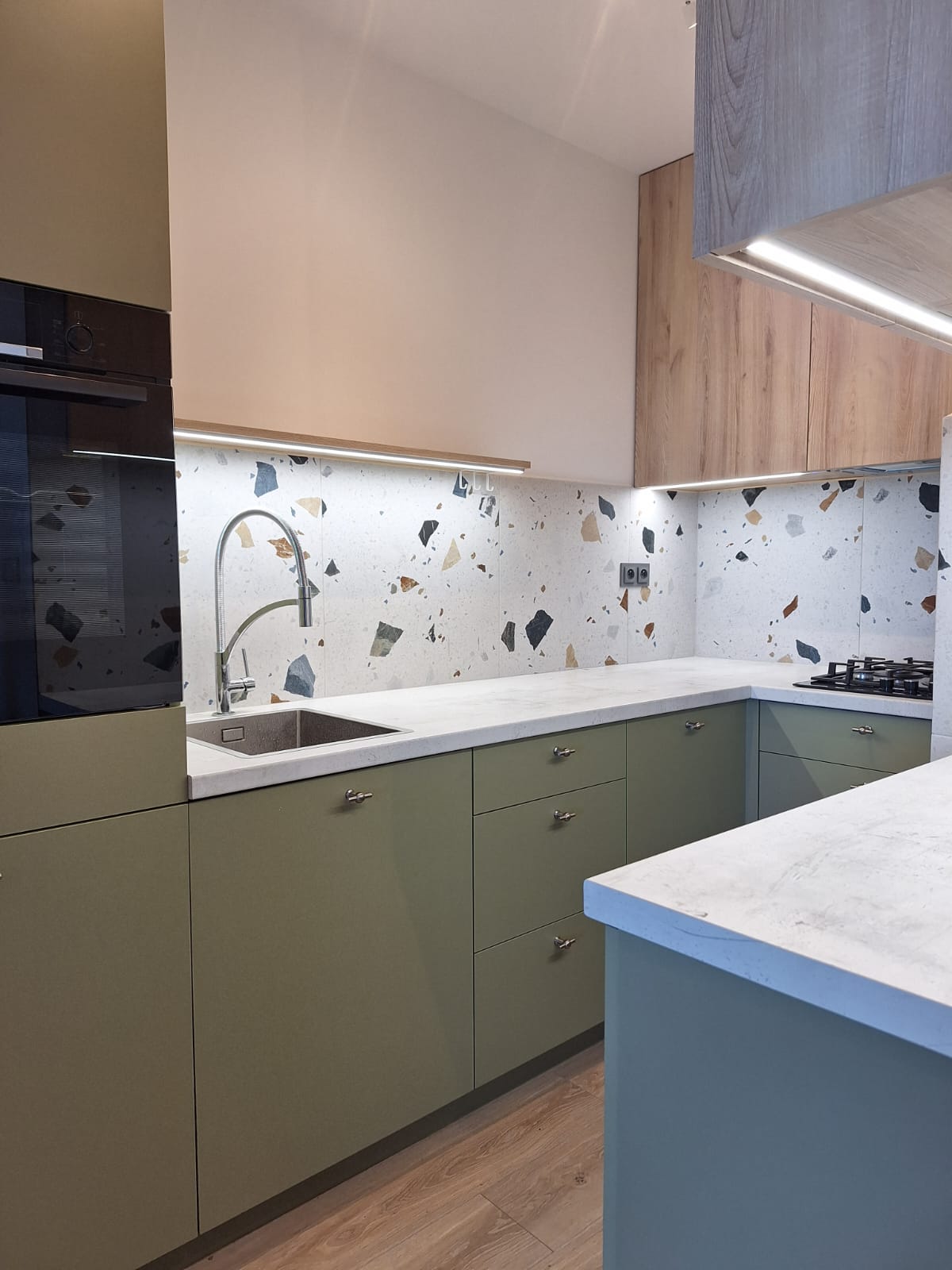 A modern kitchen is presented, featuring olive green cabinets with sleek silver handles. A white countertop extends along the area, complemented by a decorative backsplash with a mix of light-colored stones. A stainless steel sink and a built-in stovetop are visible, enhancing functionality.