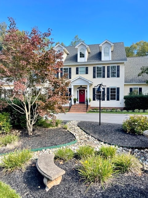 A charming two-story home is framed by lush greenery and colorful landscaping. The entry is accentuated by a bold red front door and large windows adorned with black shutters. Stone pathways and a mix of shrubs and flowers enhance the inviting curb appeal.