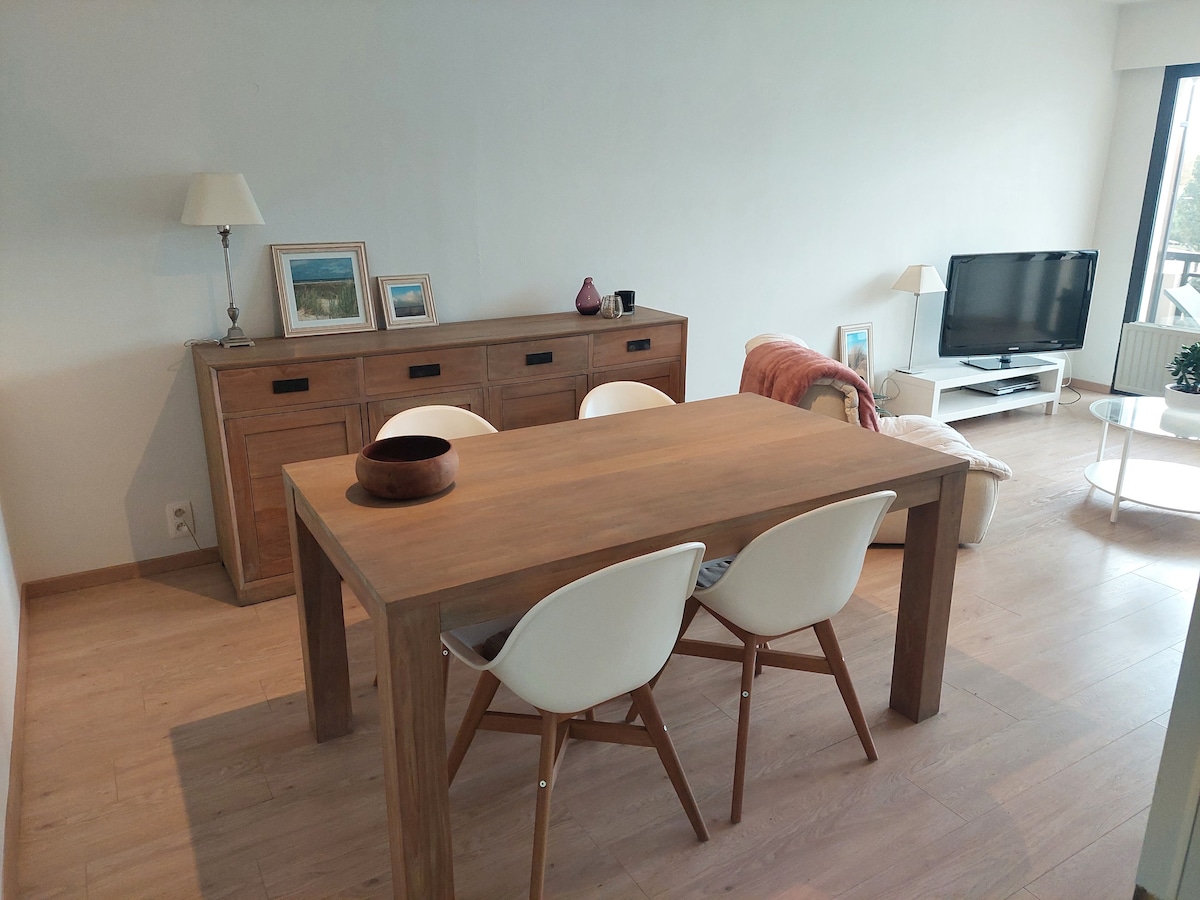 A dining area features a wooden table with three white chairs. A sideboard is positioned against the wall, displaying framed artwork and decorative items. A television is situated in the corner, complemented by a cozy armchair and natural light flowing in from the nearby window.