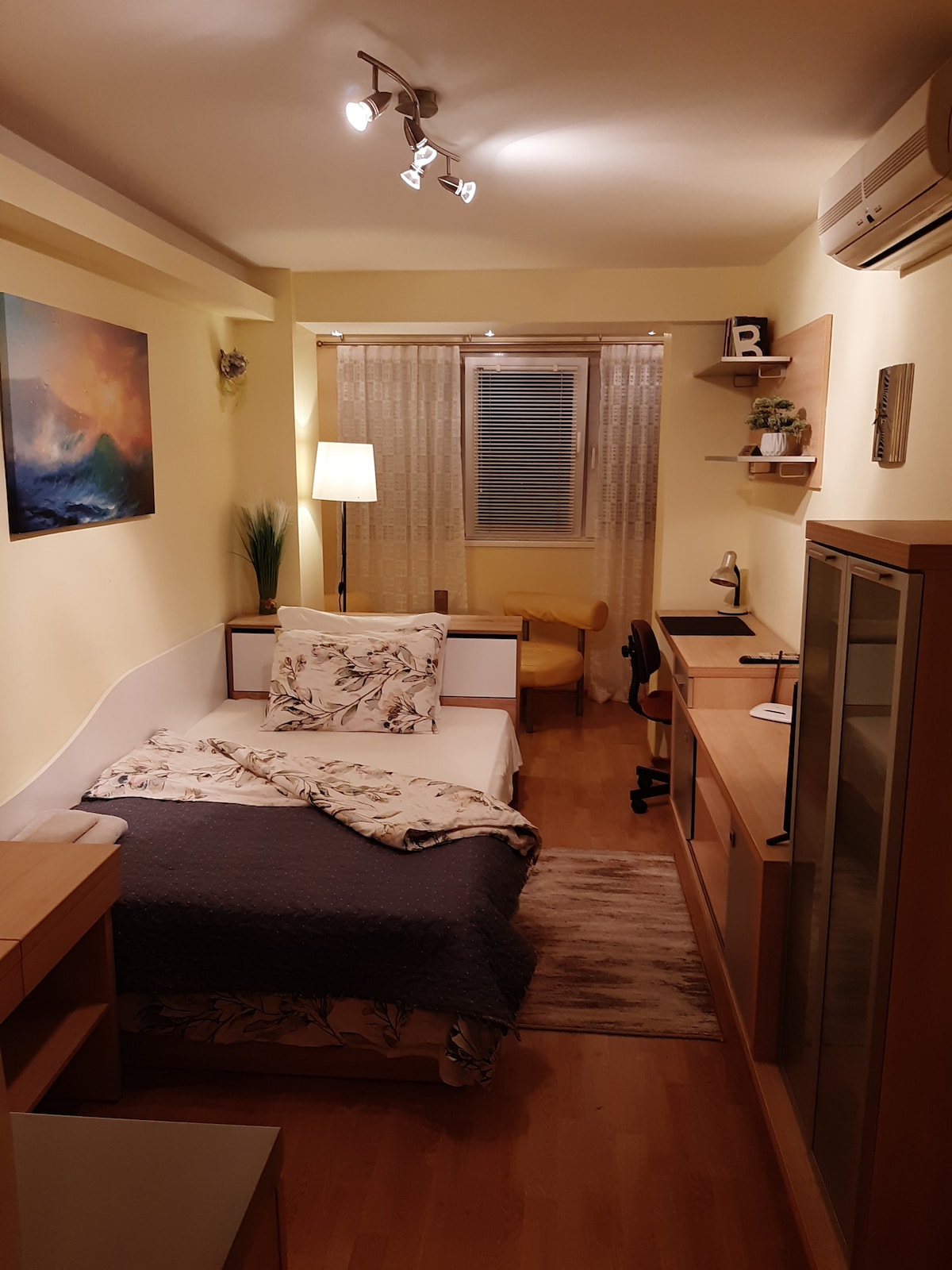 A cozy guest room features a spacious bed adorned with a floral blanket. A soft rug lies beneath, complementing wooden furniture, including a desk and shelving. Natural light filters through sheer curtains, and a small chair creates a functional seating area, enhancing the room's inviting ambiance.