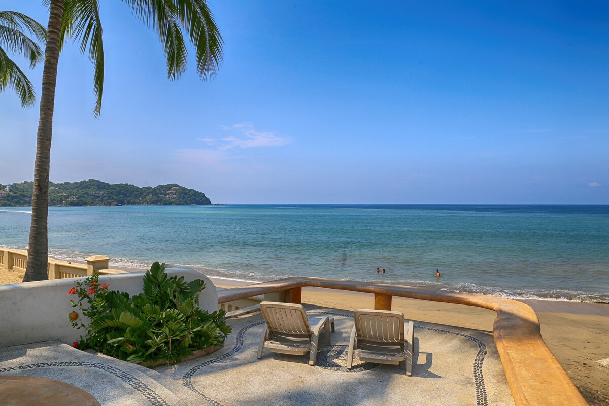 A serene beachfront view showcases two lounge chairs positioned on a stone terrace. The ocean stretches in the background, with gentle waves lapping at the shore. Lush greenery and colorful flowers frame the scene, with a distant coastline visible under a clear blue sky.